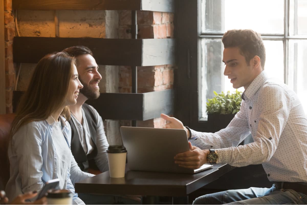 A young couple having a meeting with a young professional in a cafe setting. They appear jovial while the young professsional gestures at the laptop on the table.