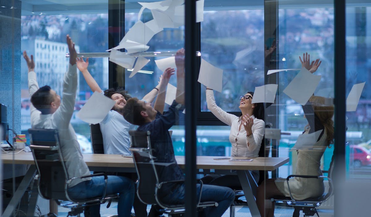 An office meeting with 5 attendees around a table throwing papers into the air